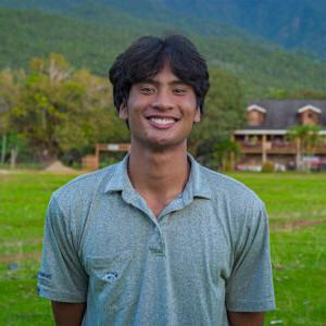 A portrait photo of Sammy Basa standing on a field of green grass with rolling green hills in the background. Basa has short dark hair and wears a gray polo shirt.